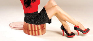 A woman in a retro polka dot skirt and red top sits on a vintage hatbox, wearing red and black polka dot pin-up heels with bows.