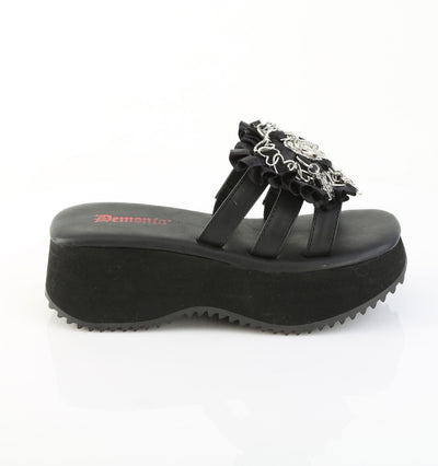 Side view of black Demonia flip-16 platform sandal with thick wedge sole, triple black straps, ruffled trim, and silver gothic heart and cross charms.
