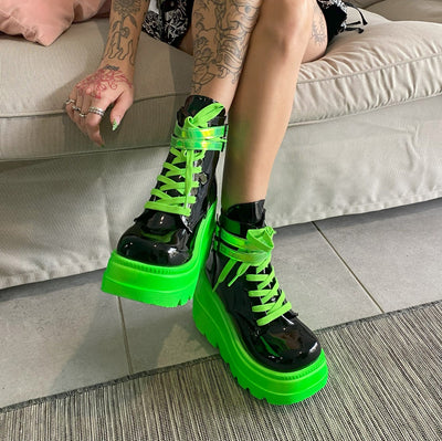 Model wearing Demonia Shaker-52 black and neon green platform boots, sitting on a couch with legs crossed.