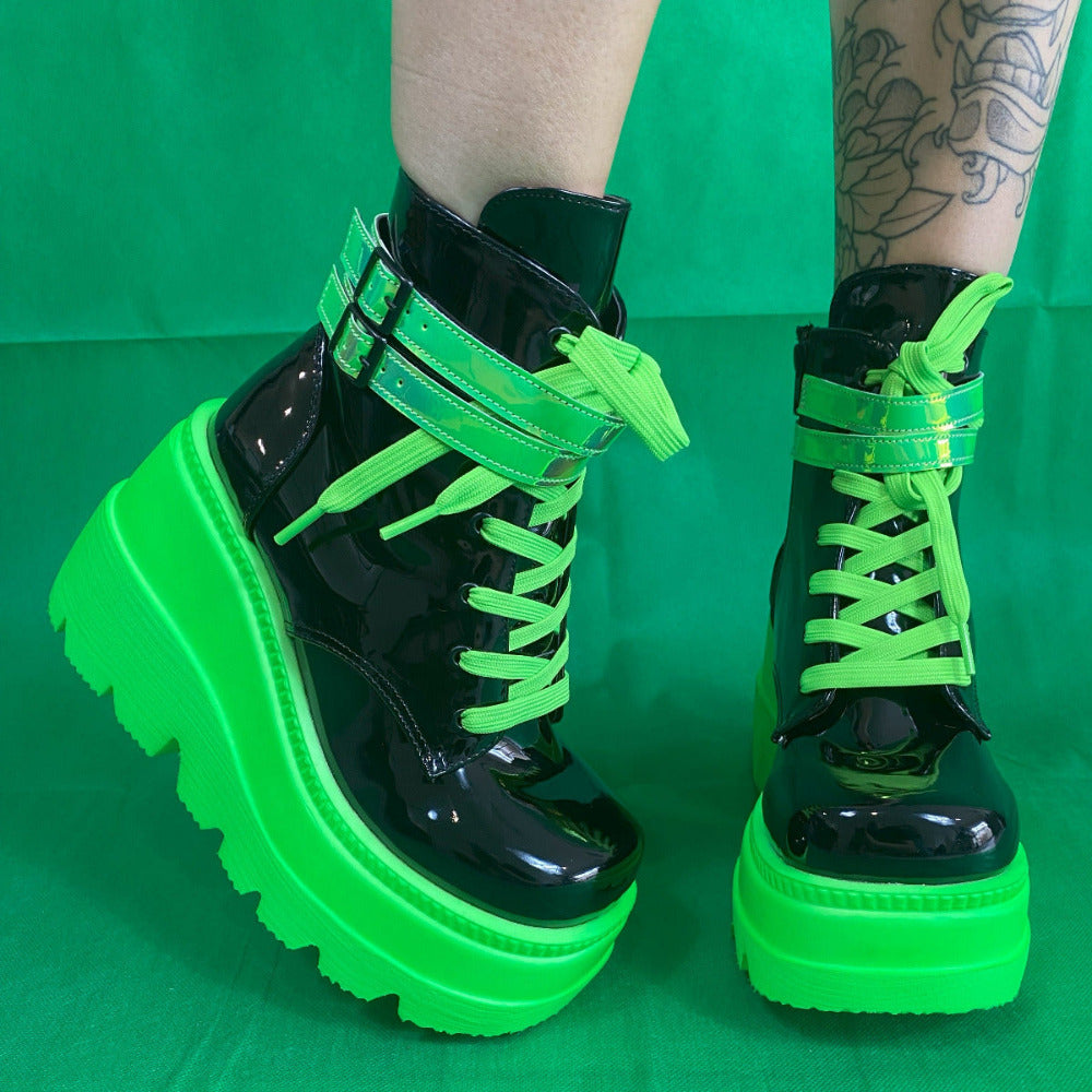 Black patent platform boots with neon green laces, straps, and sole, worn on a green background. UV-reactive style.